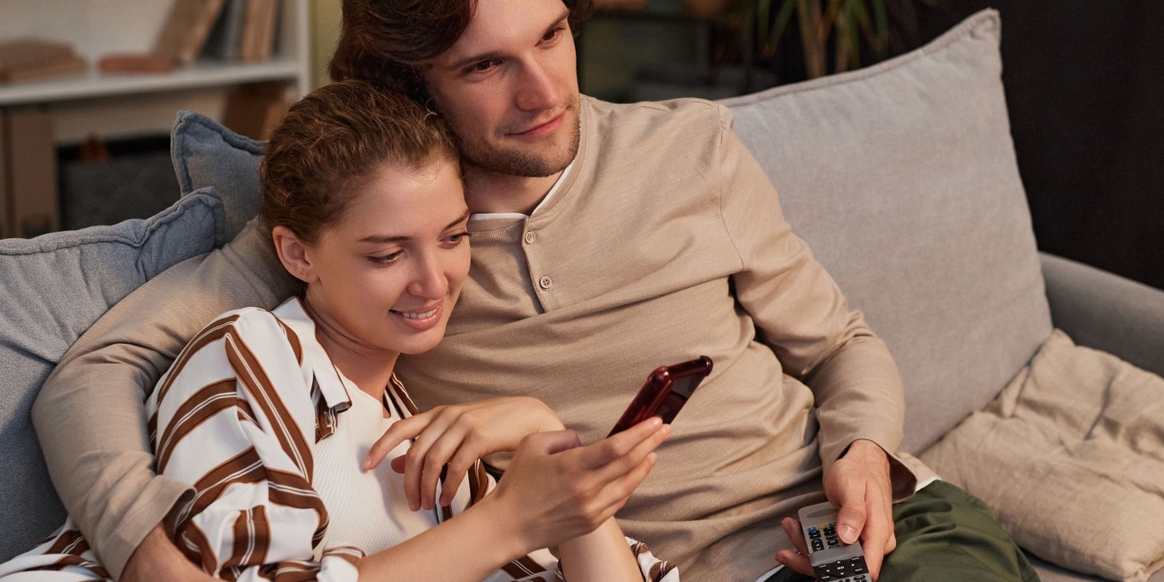A white man with short dark hair wearing a light top and dark trousers holding a TV remote control sits on a sofa with his arm round a white woman who is wearing a striped top and light jeans and is looking at a mobile phone